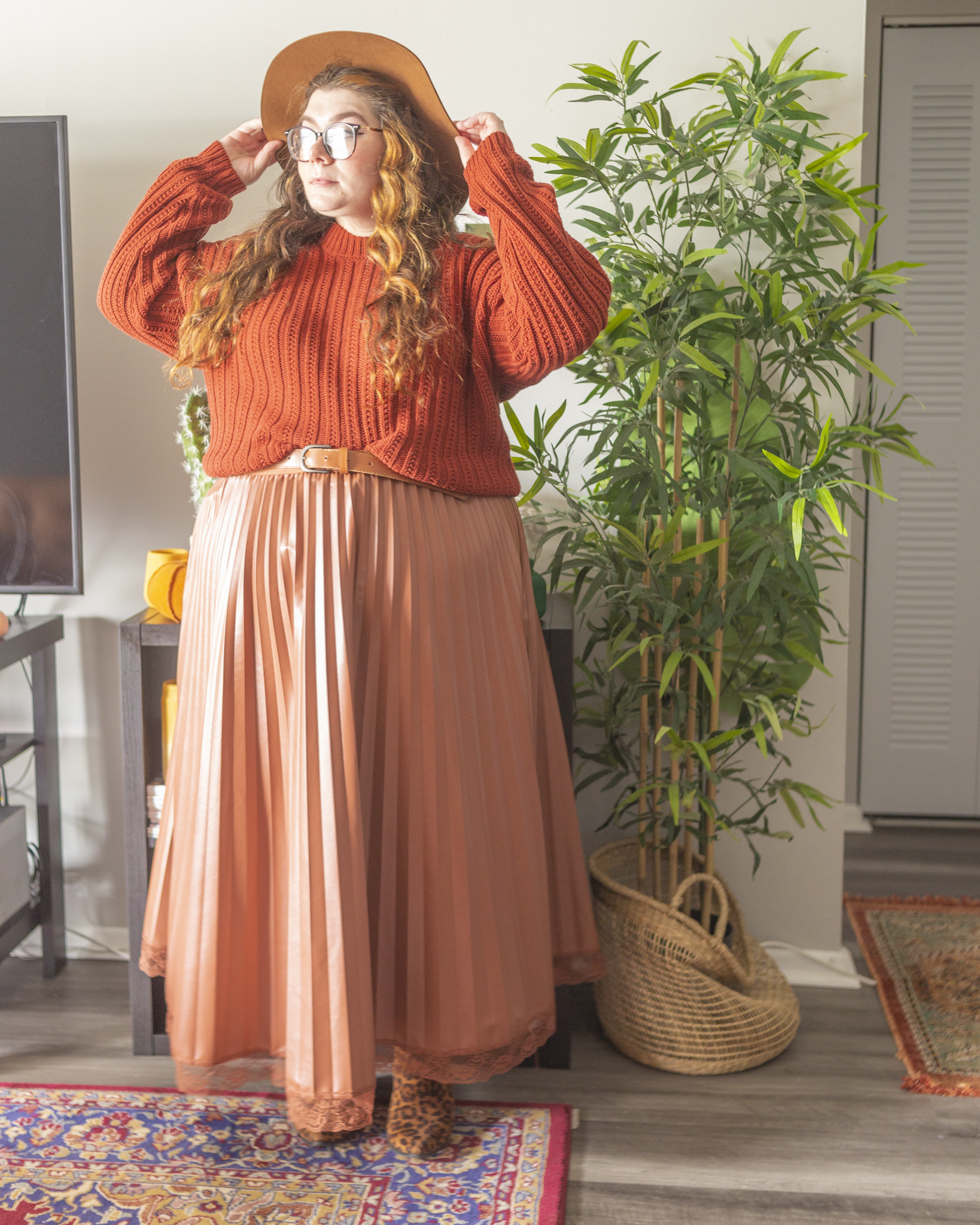 An outfit consisting of a wide brim brown hat, a dark burnt orange knit sweater half tucked into a cognac brown faux leather maxi dress with cognac brown lace trim on the bottom hem, and black on brown animal print boots.