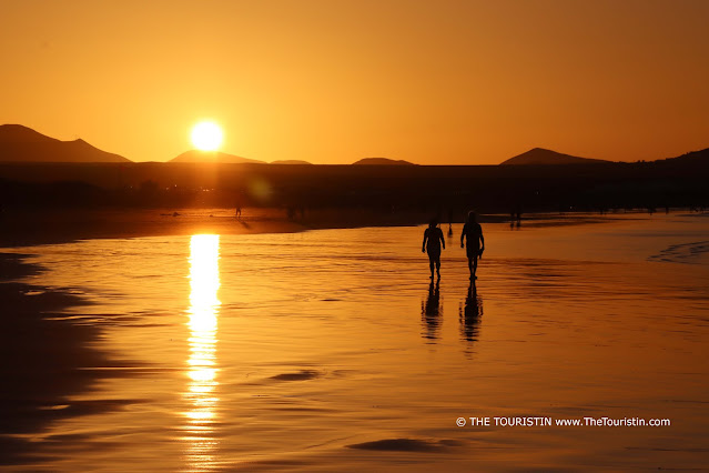 Silhouette of two beachgoers in a bright orange-red coloured landscape during sunset reflected in a thin layer of seawater on the beach. The sun sits atop a volcano crater in the distance.