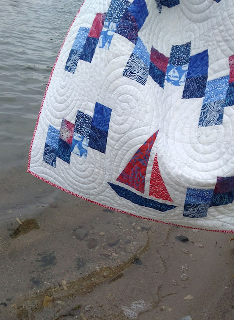 Closeup of a sailboat quilt block in a quilt, photographed  on a beach near the water.  Quilt features red and blue fabrics on a light background.