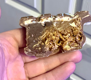 A white hand holding a rectangular piece of dark brown chocolate with a cross section showing of a light golden brown cornflake block with golden brown caramel in the middle and white salt, white chocolate spread on a bright background