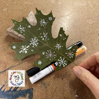 A pressed maple leaf is being held by a hand. It is covered in shiny snowflake doodles.