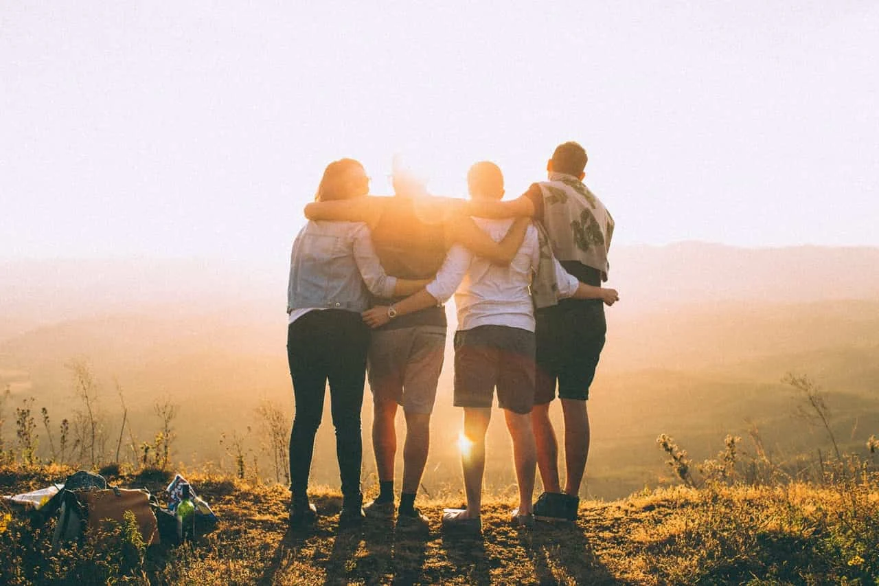 Four people standing on a cliff in front of sun hugging each other