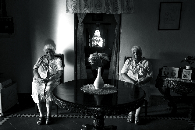 Ao centro da fotografia há uma mesa com um vaso de flor. Pouco mais ao fundo duas mulheres estão sentadas, uma do lado direito da mesa e a outra do lado esquerdo. Mais ao fundo temos uma porta, e em outro plano mais fundo uma janela, ambas com cortinas amarradas.