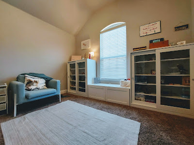 interior of a room with beige walls and a window. White cabinets with glass doors frame the window and a bench beneath the window acts as a seat. To the left is a blue armchair and on the floor is a beige throw rug.