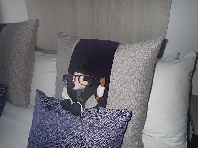 A bed made up with cushions, and a gravedigger Mickey Mouse plushie who is wearing my reading glasses.