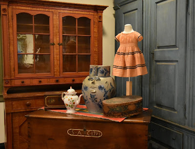 Objects from the exhibit include: a wooden blanket chest with initials "SAC 1793," a white porcelain coffee pot with floral decoration, a very large pottery jug with a stylized face and blue flowers, and a light pink child's dress displayed on a dress form