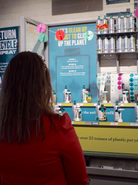 A picture from behind, showing Charlotte looking at the new bright green and blue refill station, wearing her fuchsia pink blazer.