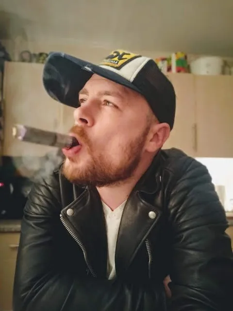 Sexy dude wearing baseball cap and a black leather jacket while smoking a cigar.