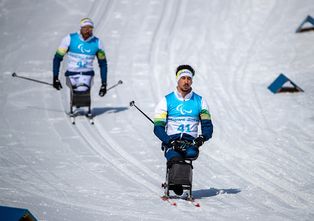 Guilherme esquia usando um par de esquis adaptados. Ele veste um agasalho azul e branco e um colete com o número 41. Robelson Lula esquia ao fundo