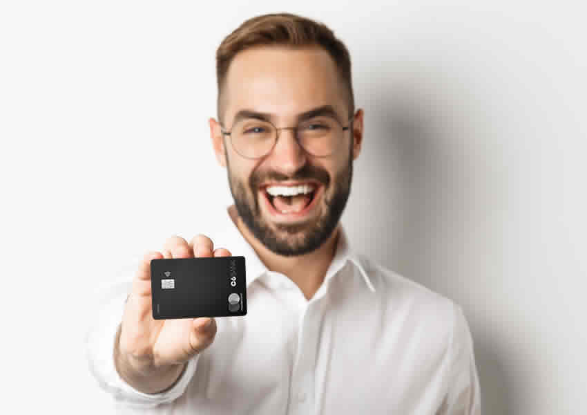 Imagem mostra um homem de óculos e camisa banca sorrindo e segurando o cartão de crédito C6 Bank Carbon sobre um fundo branco.
