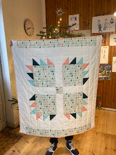 A geometric quilt being held up inside a room. you can see the feet of the person holding it. The quilt is white, peach and blue and has triangles on it. The room has a wooden floor and children's drawings on the wall. There is a christmas tree.