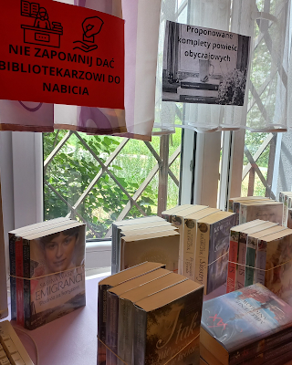 Na tle okna na firance przyczepione są dwie kartki jedna przypomina o daniu książki do nabicia bibliotekarzowi, a druga informuje, że znajdują się tutaj  komplety powieści obyczajowej proponowane przez bibliotekarza. Są one  ustawione na blacie, po dwie trzy cztery sztuki książek spiętych gumką. Jeden komplet leży, gdzie pierwszy tom nosi tytuł Milczenie aniołów. Inne tytuły są pionowo postawione, pierwsze tytuły niektórych z nich to Siostry, I tak nie przestanę Cię kochać czy Emigranci Podróż za horyzont.