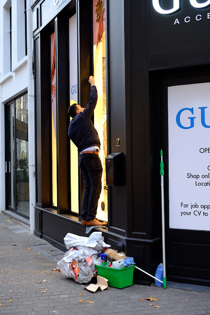 Een man, staande op de vensterbank van een winkel, is bezig met  de belettering op de winkelruit. Op de hoek ligt een stapel papier/plasticafval en er staat een grote emmer met schoonmaakproducten.