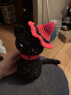 A toy cat crocheted from black with gold metallic accent yarn wearing a witch's hat over its left ear and a collar. The hat and collar are crocheted from a long color-change yarn in magenta to pink-orange. The hat's "band" and the embroidered J on the collar are in blue shiny yarn.