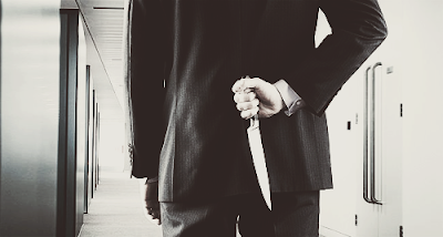 A man in a suite faces away from the camera toward a hallway with a long knife held behind his back