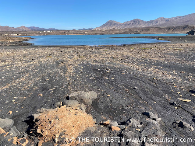 A large blue lagoon surrounded by rugged cliffs and black lava sand and rocks under a blue sky.