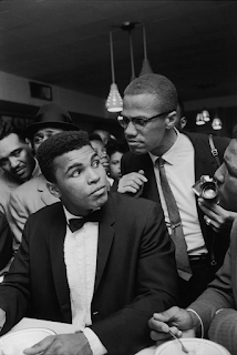 black and white photograph of Cassius Clay (Muhammad Ali) with Malcolm X leaning over his shoulder while at the victory party following his defeat of Sonny Liston, Miami, 1964