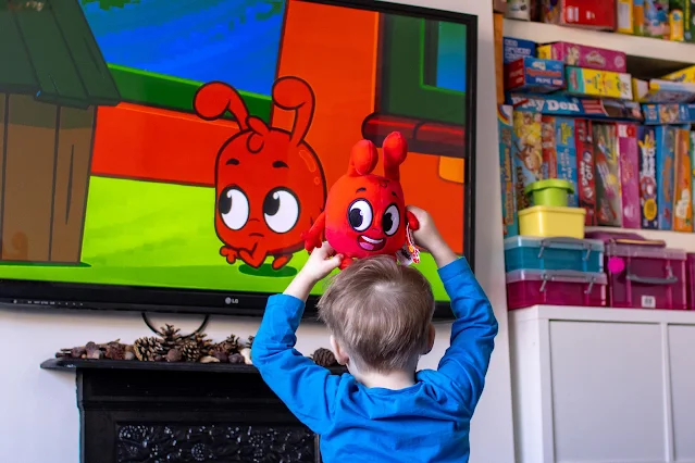 A toddler in a blue top with a red Morphle toy on his head watching an episode of Morphle on TV