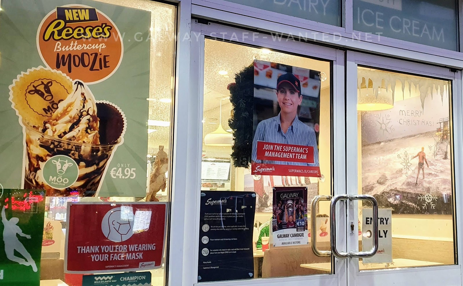 Supermacks shopfront with staff wanted sign for managers.  Also a poster for the new Reeses's buttercup moozie (ice-cream sunday) and in the background a photo of a man in bathing shorts on Salthill diving tower on a stormy day