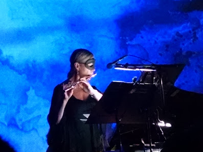 A woman in a black dress and black half mask plays the flute in front of a projection resembling the sea. The flutist's music stand, a microphone, and a piano are also visible in the photo.