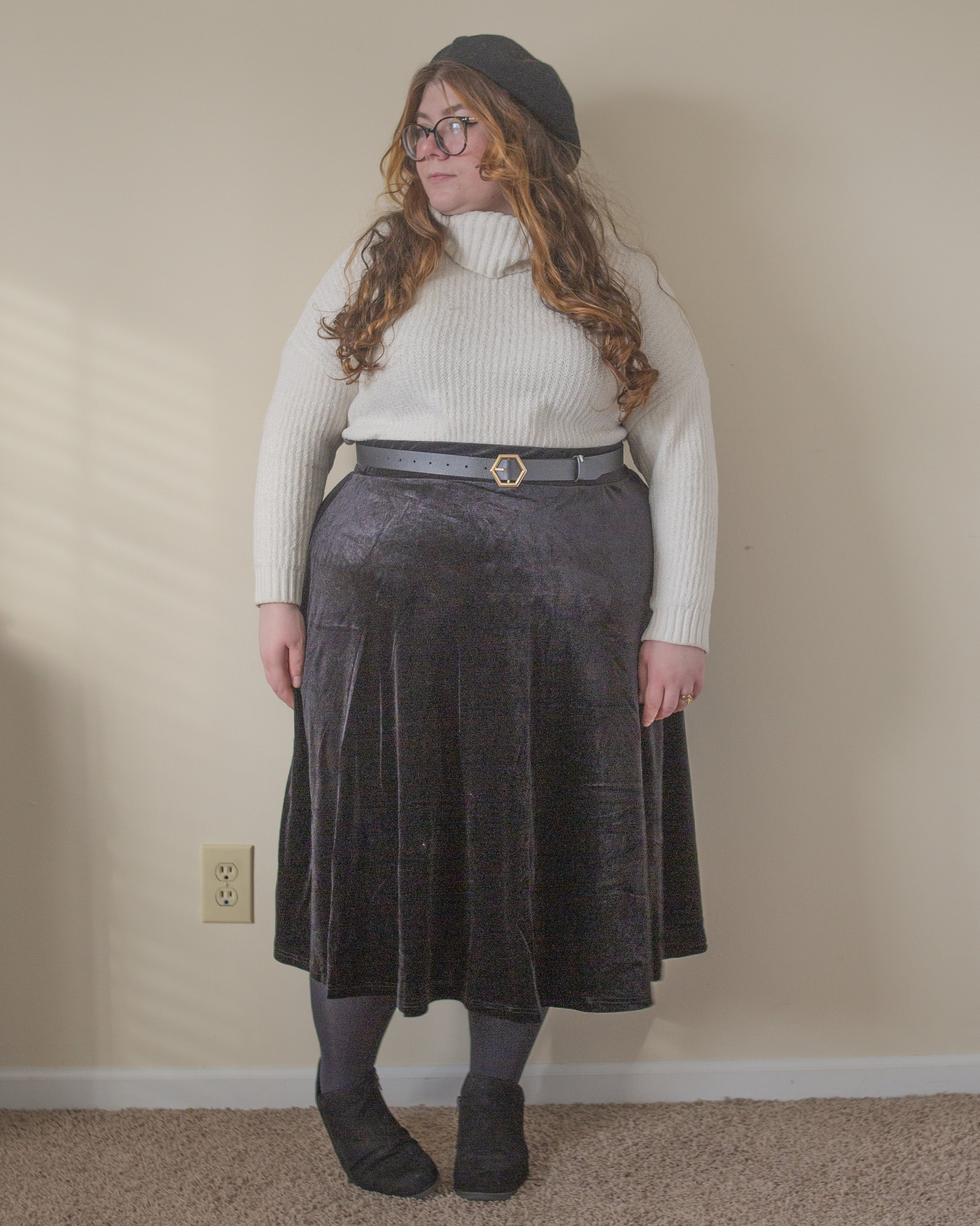 An outfit consisting of a white chenille turtleneck half tucked into a black velvet midi skater skirt, black tights, and black ankle strap heels.