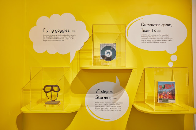 A pair of flying goggles, a vinyl record, and the 1990s computer game Worms in individual display cases with accompanying text