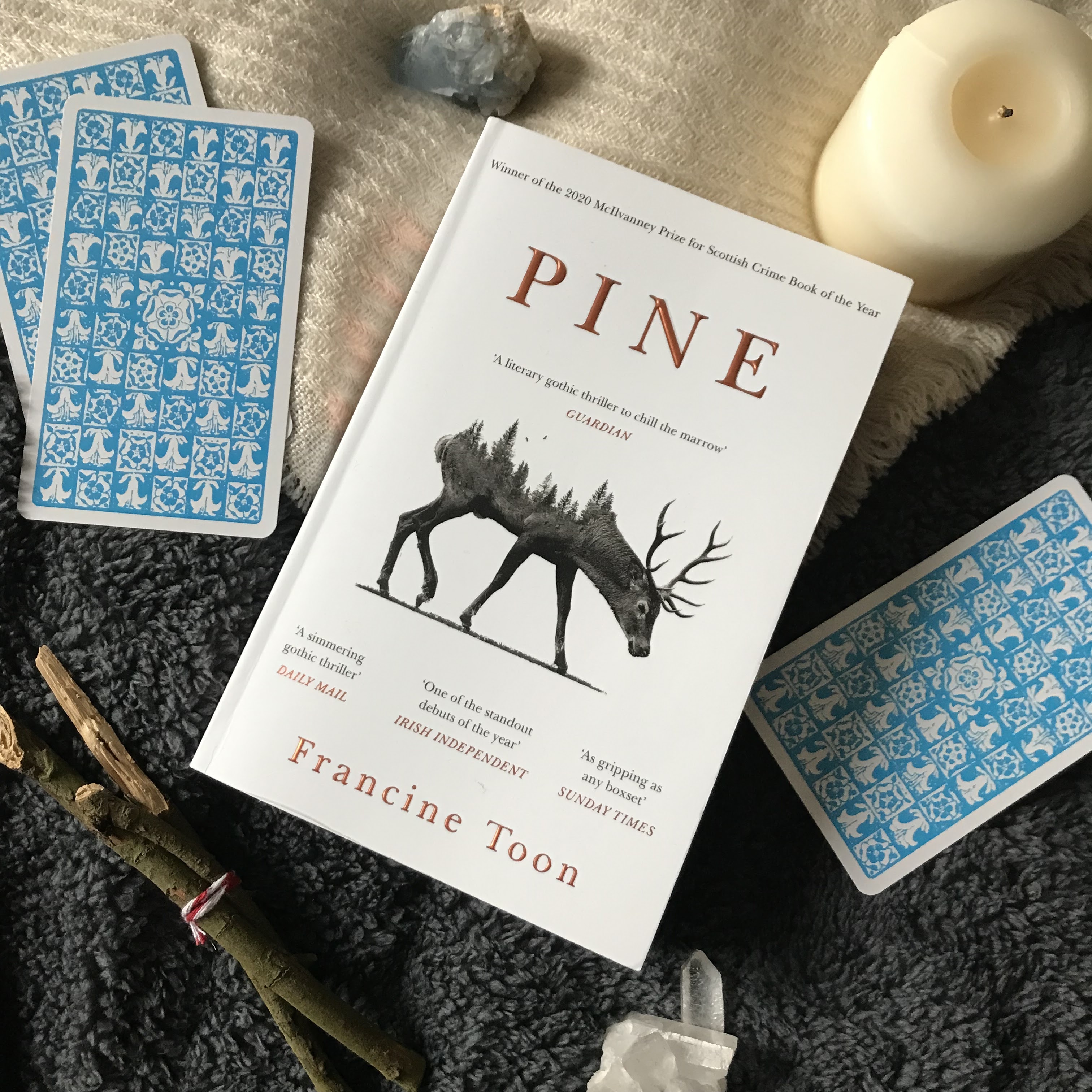 A photo of Pine by Francine Toon, half on a wool cream scarf, half on a dark grey, fluffy duvet cover. The book is at an angle so the top of the book faces the top right corner, and the bottom faces the bottom left corner. The book is surrounded by various objects; in a clockwise direction from the top right, there's a pillar candle, a single tarot card, face down, a clear quartz crystal cluster, a bundle of yarrow twigs, two tarot cards, face down, overlapping each other, and a rough blue calcite crystal.