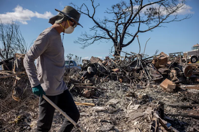 man in protective face mask clears debris of destroyed house where wildfires burned in Maui