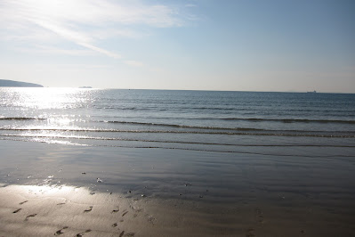 The beach and sea at broadhaven. The sun is reflecting off the water on the left side of the image. The sky is blue with a little bit of white cloud.