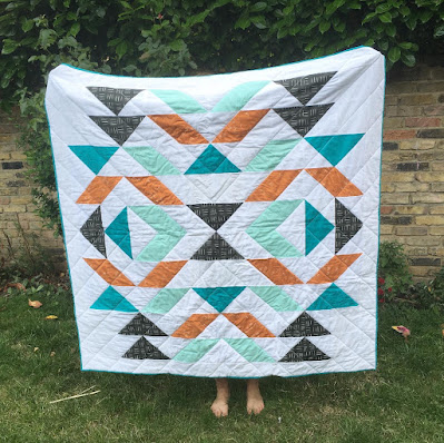 Large quilt with geometric shapes in blues, terracotta and black on a white background. The quilt is being held up by a person (unseen) in a garden, with a brick wall behind.