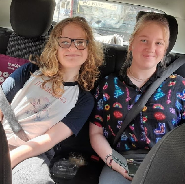 Will on the left wears a white and black t shirt and leans in next to L wearing jeans and a blue funky shirt. The are wearing seatbelts in the back of the car and smiling