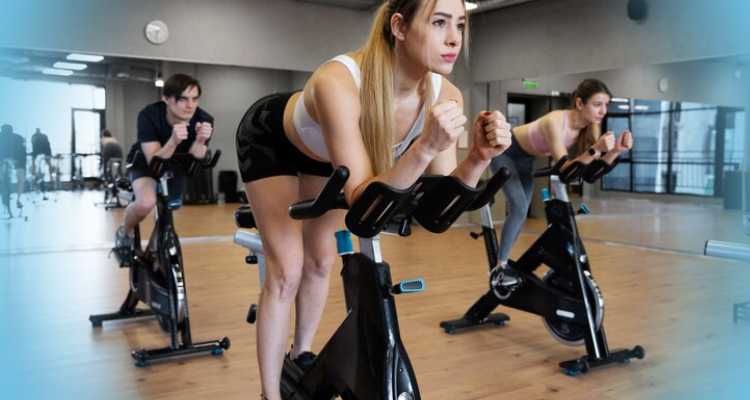 a woman in a gym, using an indoor cycling stationary bicycle. She is wearing workout clothes and footwear, with her knee and joint visible. The background includes a wall and gym floor.