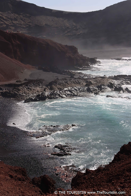 Waves crash on a black lava beach surrounded by red cliffs; silky droplets of ocean spray fills the air.