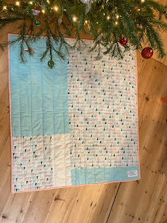 A quilt laid out on a wooden floor. The quilt is white, peach and blue and has triangles on it. There is a christmas tree visible in the top of the frame.