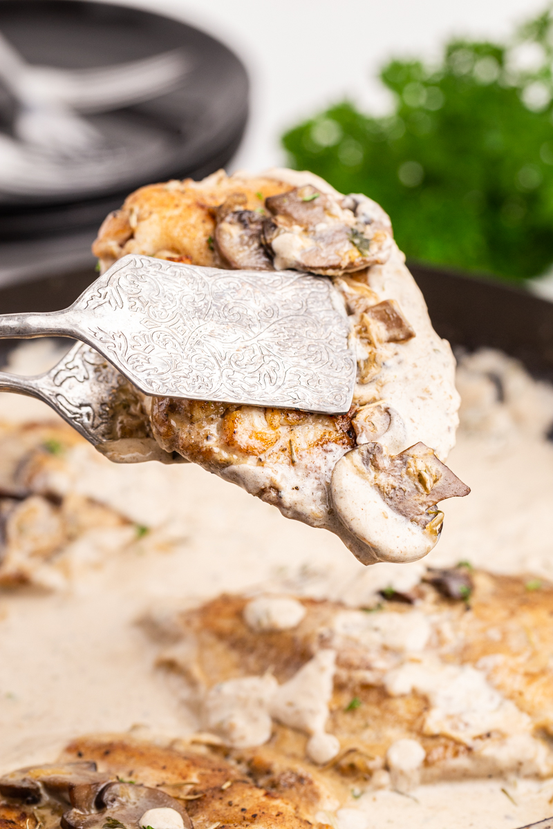 Closeup photo of one piece of Chicken with Mushroom Garlic Sauce being held with silver tongs above the skillet.