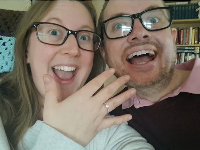 Hannah on the left smiling opened mouthed wearing glasses shows off her ring on her hand next to Martyn wearing a maroon jumper and smiling widely wearing black framed glasses