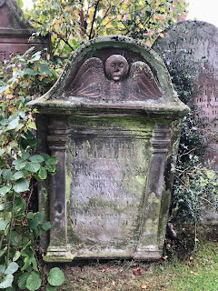 A photograph of an old gravestone in Penpont Graveyard.  At the top is carved a round face with wings underneath.  The writing on the stone is illegible.