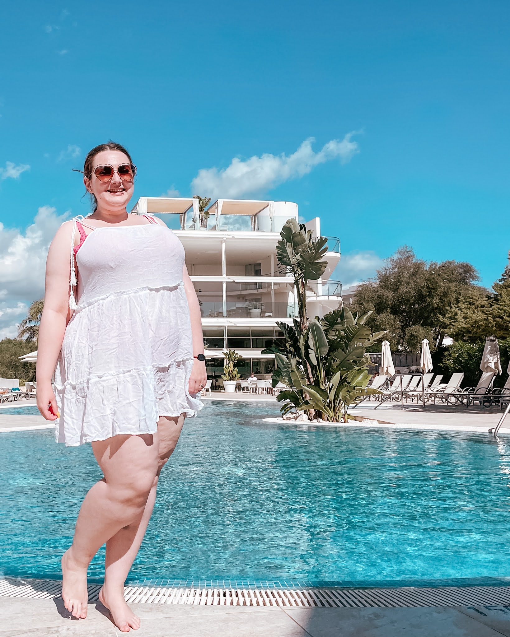 Becky wears a short white beach dress over a bright pink bikini. She stands at the edge of the bright blue swimming pool. In the background is the gleaming white hotel building