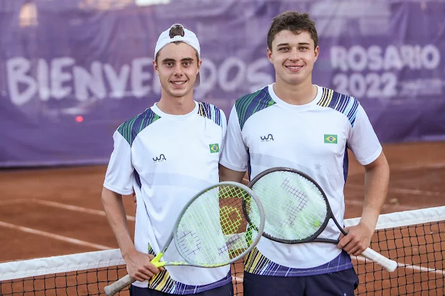 Victor e João posam para foto. Ambos vestem o uniforme branco da seleção brasileira de tênis. Ao fundo, uma quadra de saibro