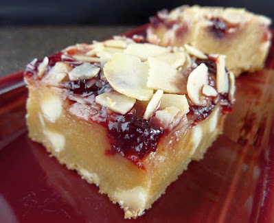 Photo of a Bakewell Blondie: a cookie base with white chocolate chips, topped with cherry jam and sliced almonds. Photographed on a burgundy plate.