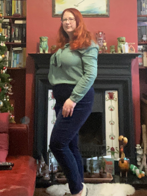 A red headed woman in slim-fitting navy velvet jeans and a sage green pussybow blouse. She is standing in front of a fireplace, next to a Christmas tree.