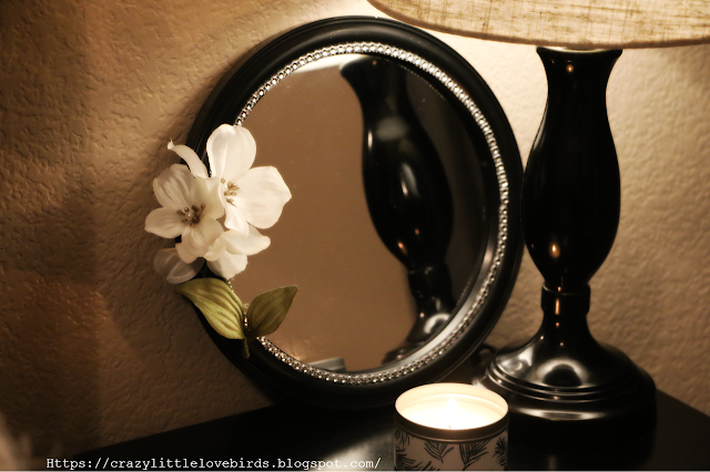 Black round mirror with faux white flowers and gems displayed near a lamp on a table