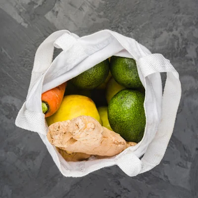 Top-down view of white reusable shopping bag containing fresh produce including avocados, lemon, carrot, and ginger root on gray surface