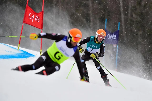 Bartagnolli esquia em uma pista de neve com bandeiras vermelhas e azuis. Ele está no fundo usando um colete azul. Na sua frente está seu atleta guia vestindo um colete amarelo com a letra G