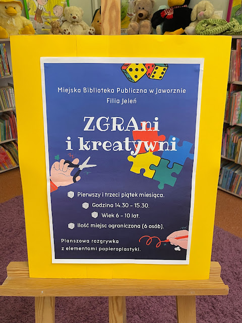 Na sztaludze znajduje się pionowy plakat, informujący o popołudniowych zajęciach dla dzieci. Na granatowym tle znajdują się białe napisy: Miejska Biblioteka Publiczna w Jaworznie - Filia Jeleń, ZGRAni i kreatywni, Godzina 14.30 - 15.30, Wiek 6 - 10 lat, Ilość miejsc ograniczona (6 osób), Planszowa rozgrywka z elementami papieroplastyki. Na plakacie dodatkowo 2 kostki do gry,  4 elementy puzzli, dłoń trzymająca ołówek, dłoń trzymająca kredkę. Sztaluga stoi na fioletowym dywanie, na tle regałów z książkami dla dzieci, na regałach siedzą poustawiane pluszaki.