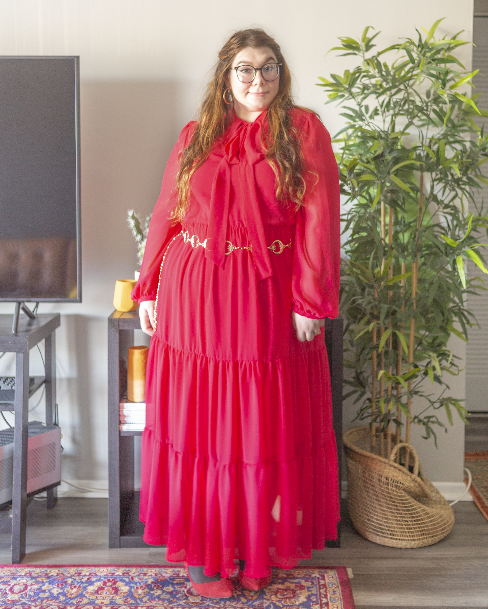 An outfit consisting of a red bishop sleeve sheer red maxi dress with an attached pussy bow, with a gold star and moon chain belt around the waist and red ankle strap heels.