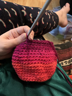 What looks like a small crocheted bag in a variegated yarn that flows from orange to orange-pink to red purple from bottom to the round being worked on.