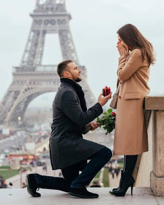Casal em Paris, ele, ajoelhado oferta uma flor para ela. #PraCegoVer