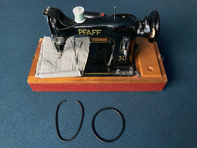 A Pfaff 30 sewing machine on the floor, with two rubber drive belts in front of it: The old broken one, and the new one, ready to be fitted.