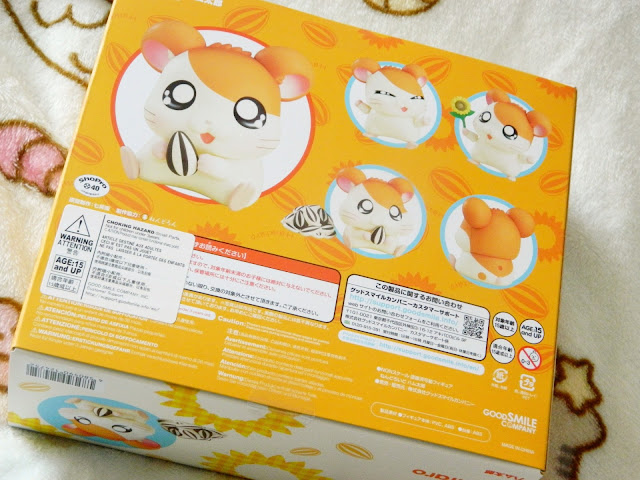 A photo showing the outer box of an Hamtaro Nendoroid figure. Hamtaro is an orange and white hamster from an anime series. The box is bright orange, and there are picture of Hamtaro all around.